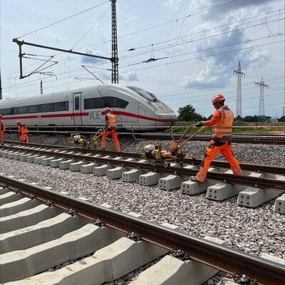 Während Personen auf einer Gleisbaustelle arbeiten, fährt im Hintergrund ein ICE vorbei.